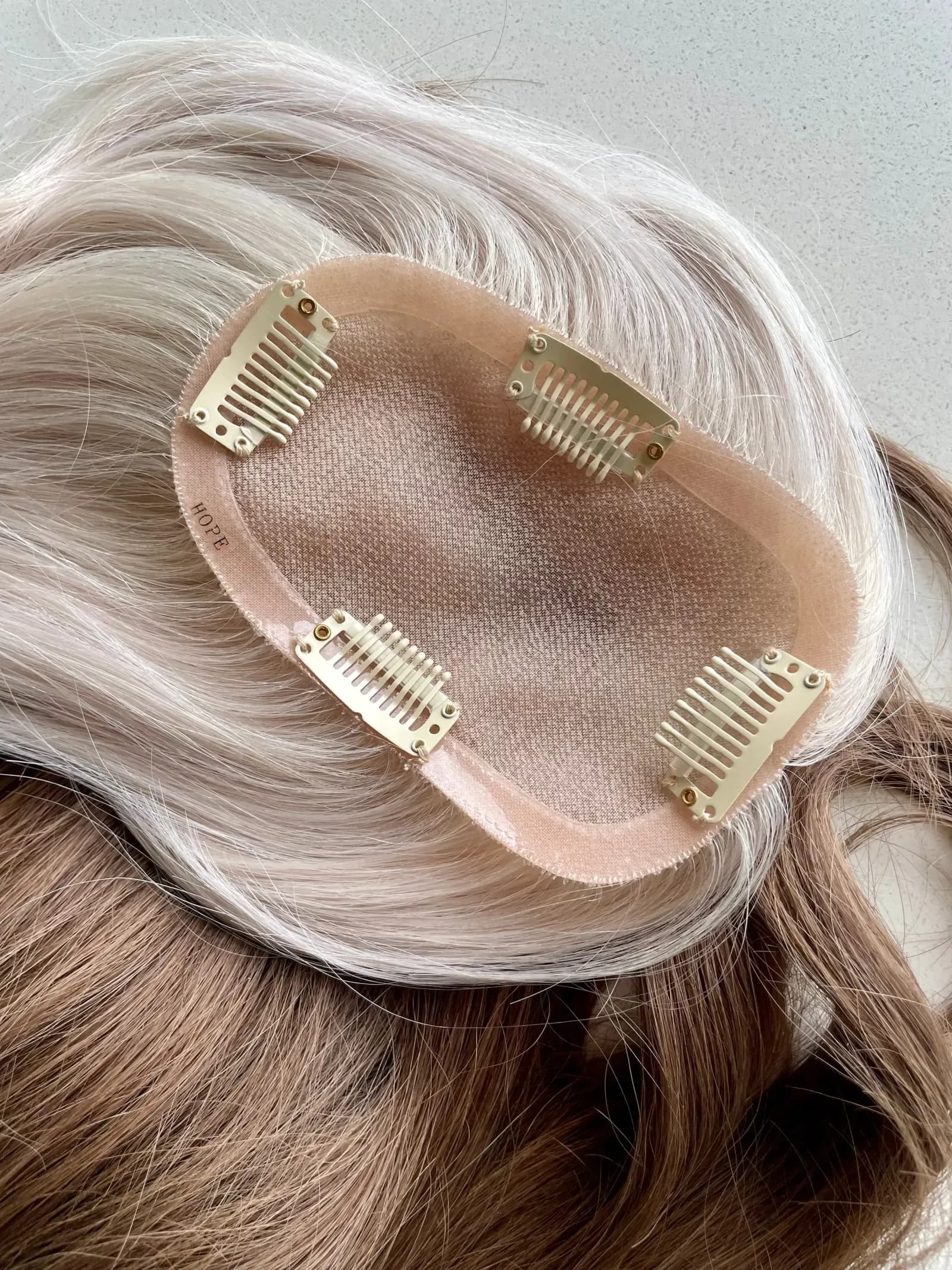Underside of a Remy human hair topper showing its silk base and four snap-clip attachments, with blonde hair fading to darker roots