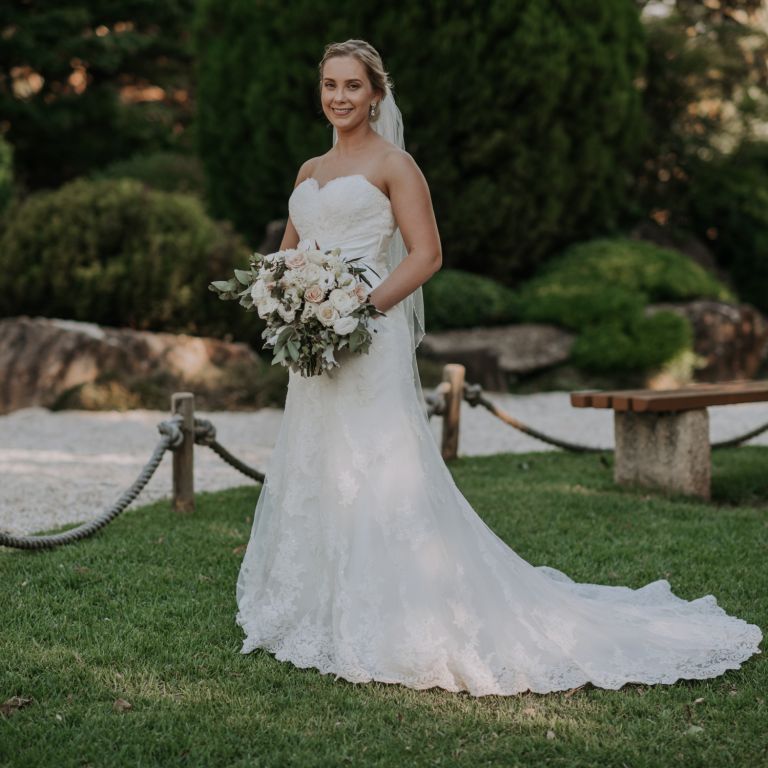 A woman in white gown and veil, holding a white bouquet of flowers, full shot