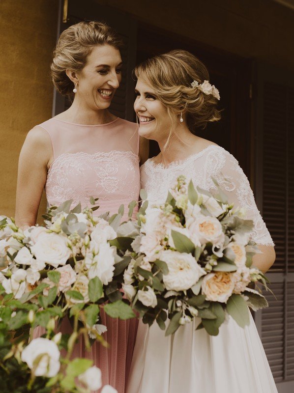 A woman in pink gown and a woman in white gown holding a set of flowers, smiling candidly